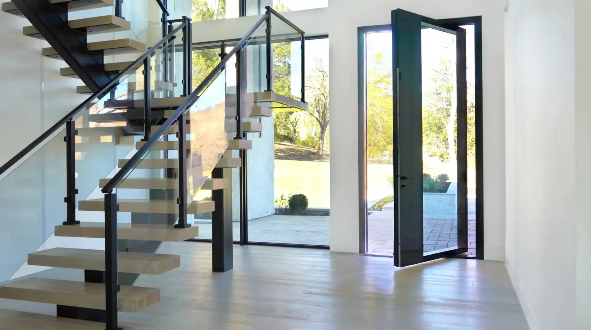 pivot_door_interior_open_scaled_c1x40c9smcn2deabmyn0l_1920x1073_8j2n2bzdbpg2gtb3p4jjgo-1920x1073 Modern entryway with floating staircase and glass railing.